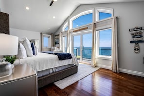 Une chambre avec un grand lit, une table de chevet, une lampe, une plante et une vue sur la mer grâce à de grandes fenêtres.