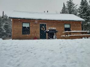 Une cabane en bois avec un toit enneigé, une porte d’entrée et un petit porche.