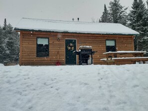 Uma cabana de madeira com telhado coberto de neve, porta da frente e um pequeno alpendre.