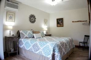 A bedroom with a bed, two bedside tables with lamps, a chair, a wall-mounted heater, and a picture frame.