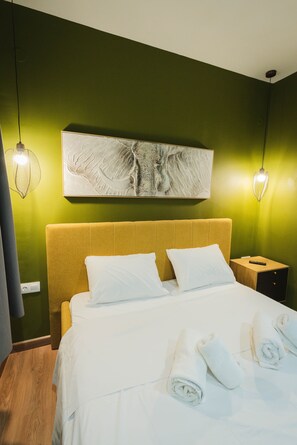 Hotellihuone, jossa on sänky, kaksi valkoista tyynyä, sängynpääty, yöpöytä, seinälle ripustettu taideteos ja kaksi riippuvalaisinta.