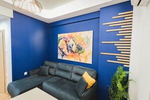 Ein Zimmer mit blauer Wand, einem schwarzen Sofa mit gelbem Kissen, einem Gemälde und Holzlatten an der Wand.
