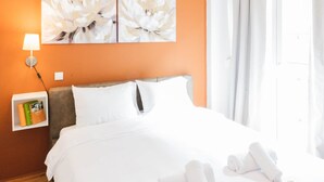 Una camera d'albergo con una parete arancione, un letto grande con lenzuola bianche, una lampada da comodino e due quadri di fiori appesi al muro.
