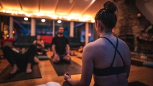 Una clase de yoga en una habitación con suelo de madera, pared de piedra y un ventanal grande.