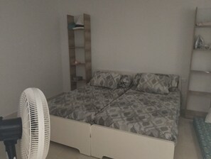 Ein Schlafzimmer mit einem Bett, einem Standventilator, einem Regal mit Gegenständen und einer Wandablage.