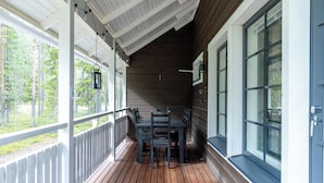 A covered porch with a wooden table and chairs, large windows, and a view of a forest.
