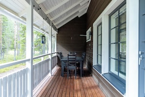 A covered porch with a wooden table and chairs, large windows, and a view of a forest.