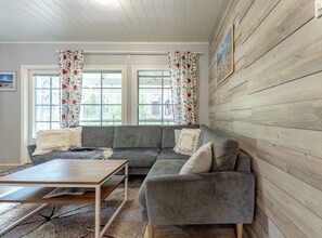 A living room with a grey sofa, a wooden coffee table, and a rug. There are two windows with floral curtains.