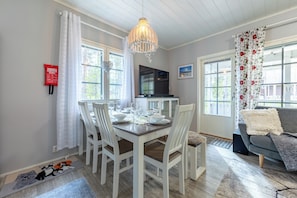 A dining area with a table set for four, white chairs, a chandelier, a TV, and a sofa in the background.