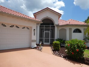 A residential house with a garage, a front door, and a small garden with plants and a bird. - Beautiful villa in SW - Cape Coral in central location within walking distance to everything (Cape Coral)