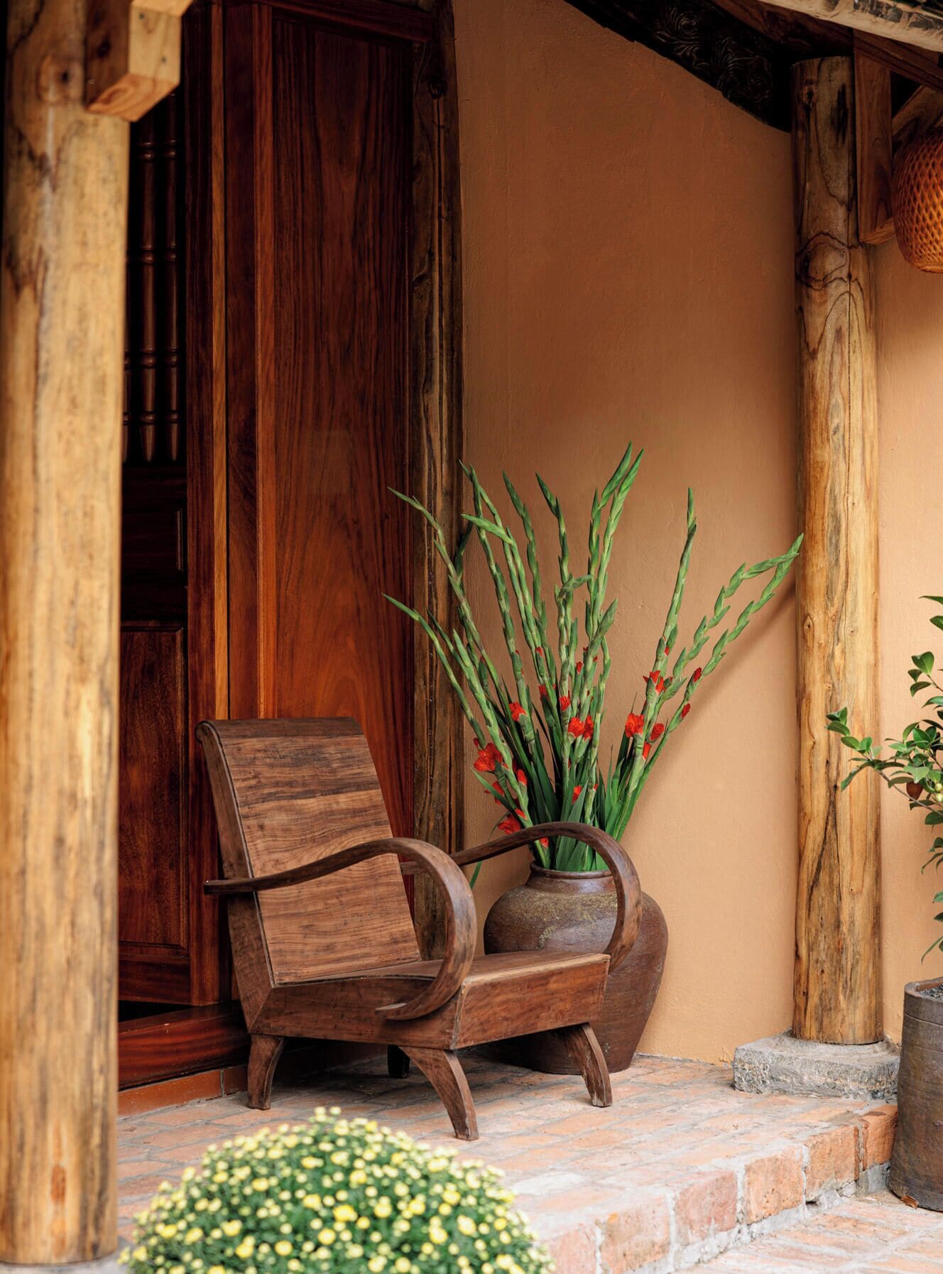 A wooden chair and a vase with plants and red flowers on a stone-paved porch.