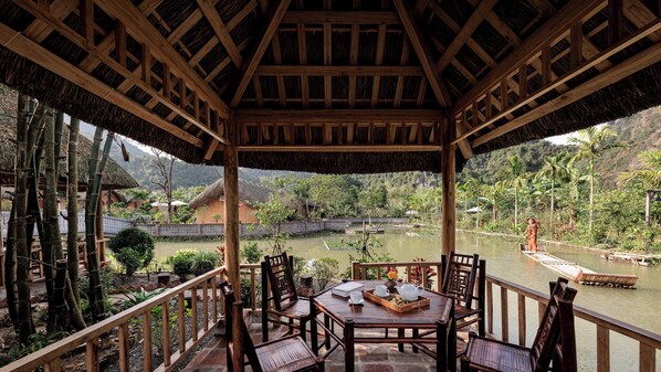 A wooden gazebo with a thatched roof overlooking a calm river, surrounded by lush greenery and traditional buildings. - The Boi Heritage Tam Coc (Hoa Lu)