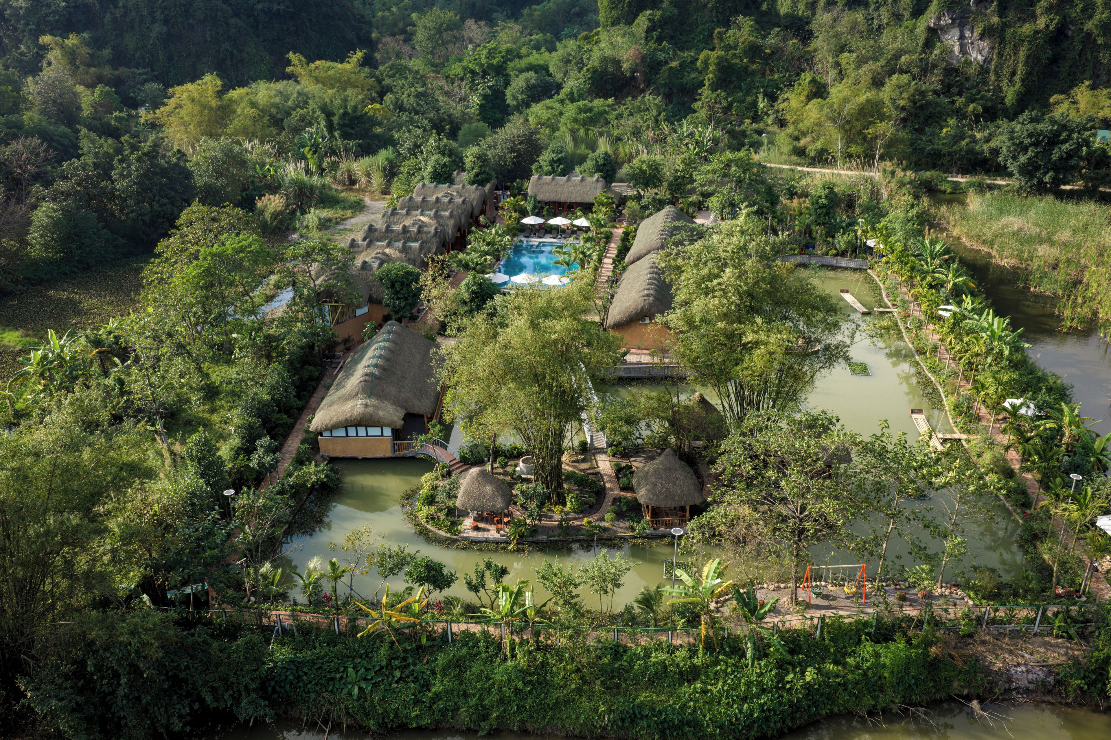 Um resort com construções de telhado de palha, uma piscina e paisagem natural ao redor.