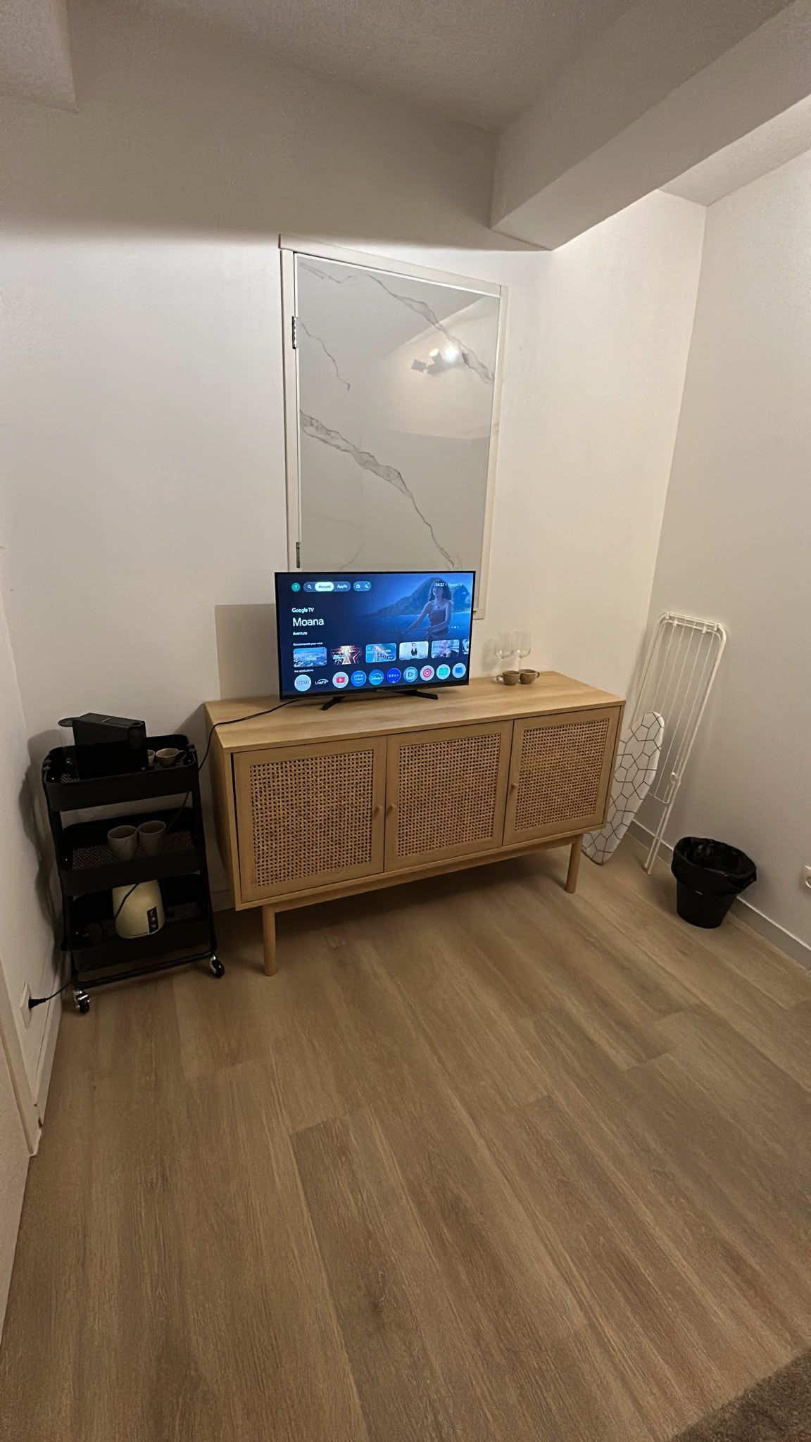 Ein Zimmer mit einem Holzkabinent, einem Fernseher mit Google TV Oberfläche, einer weißen Heizung und einer schwarzen Mülltonne.