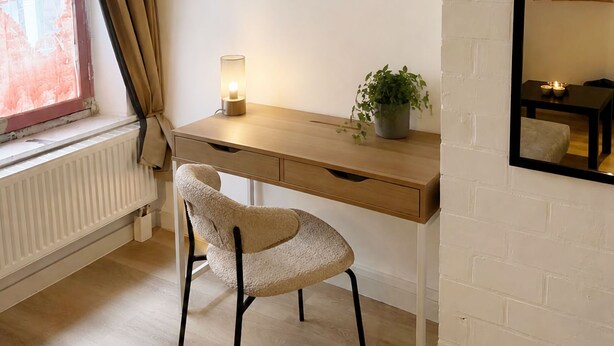 Una habitación con un escritorio de madera, una silla, un radiador, una ventana con cortinas, un espejo montado en la pared y una obra de arte en la pared.