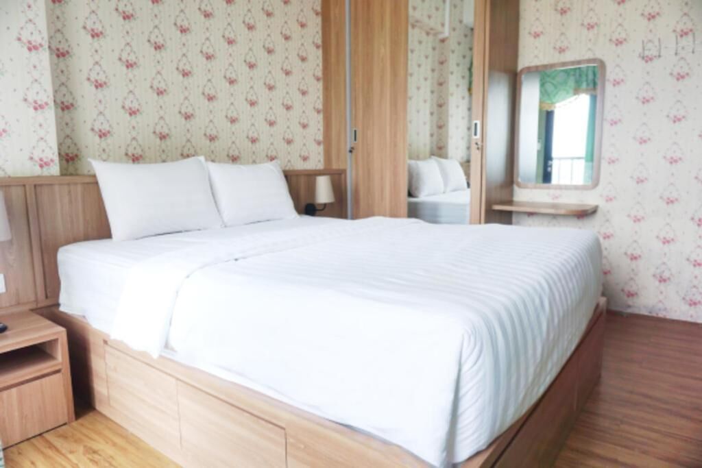 Een netjes opgemaakt bed in een kamer met bloemenbehang, een houten nachtkastje en een spiegel aan de muur.