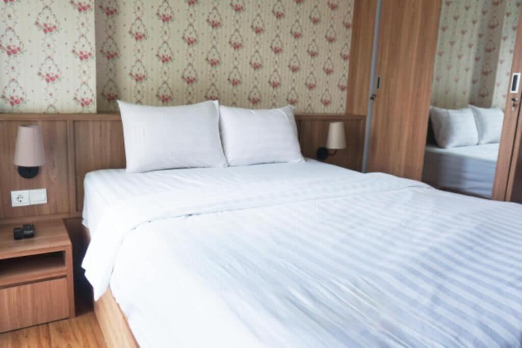 Un letto rifatto con lenzuola bianche, una testiera in legno, un comodino con una lampada e una parete con carta da parati a motivi floreali.