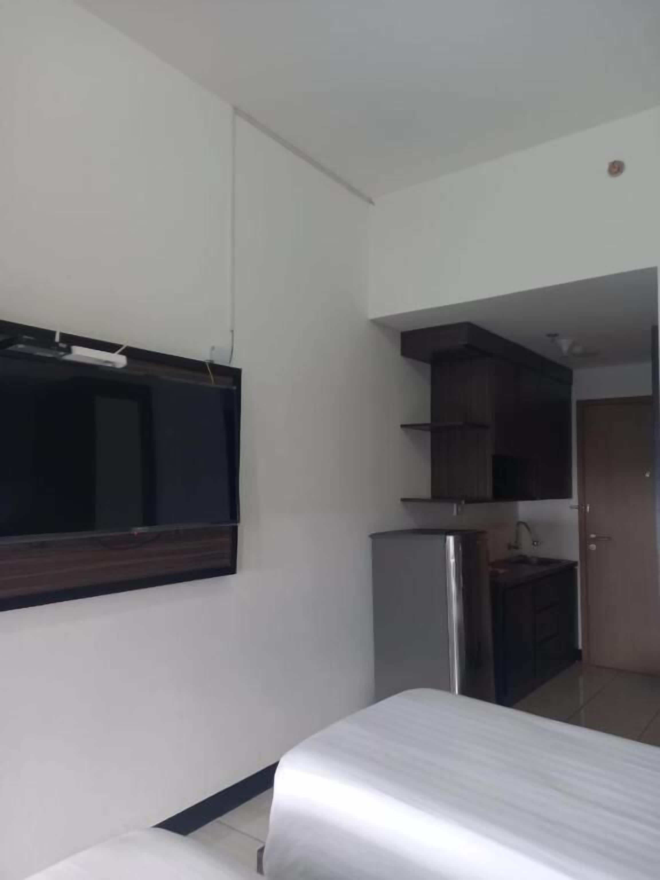 A bedroom with a bed, a TV mounted on the wall, a kitchenette with a fridge, and a door leading to another room.