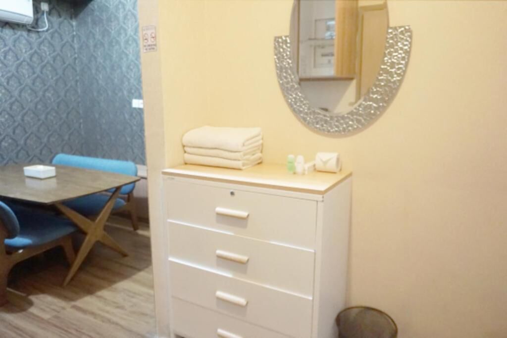 Una habitación con una mesa de madera, sillas azules, un armario blanco con cajones, un espejo y un cubo de basura pequeño.