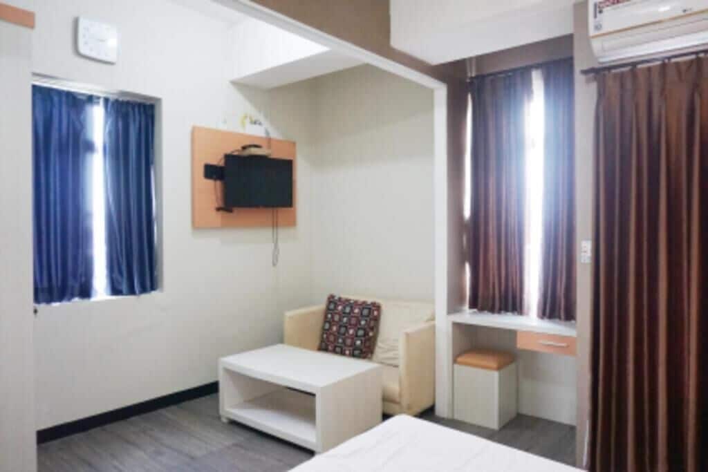 A hotel room with a bed, a small white table, a sofa, a TV mounted on the wall, and a window with curtains.