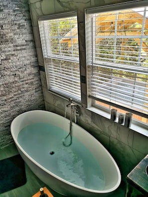 A bathroom with a freestanding bathtub, two windows with blinds, and a view of a wooden structure outside. - Romantic Granbury Cabin Ideal for a Texas Getaway (Granbury)
