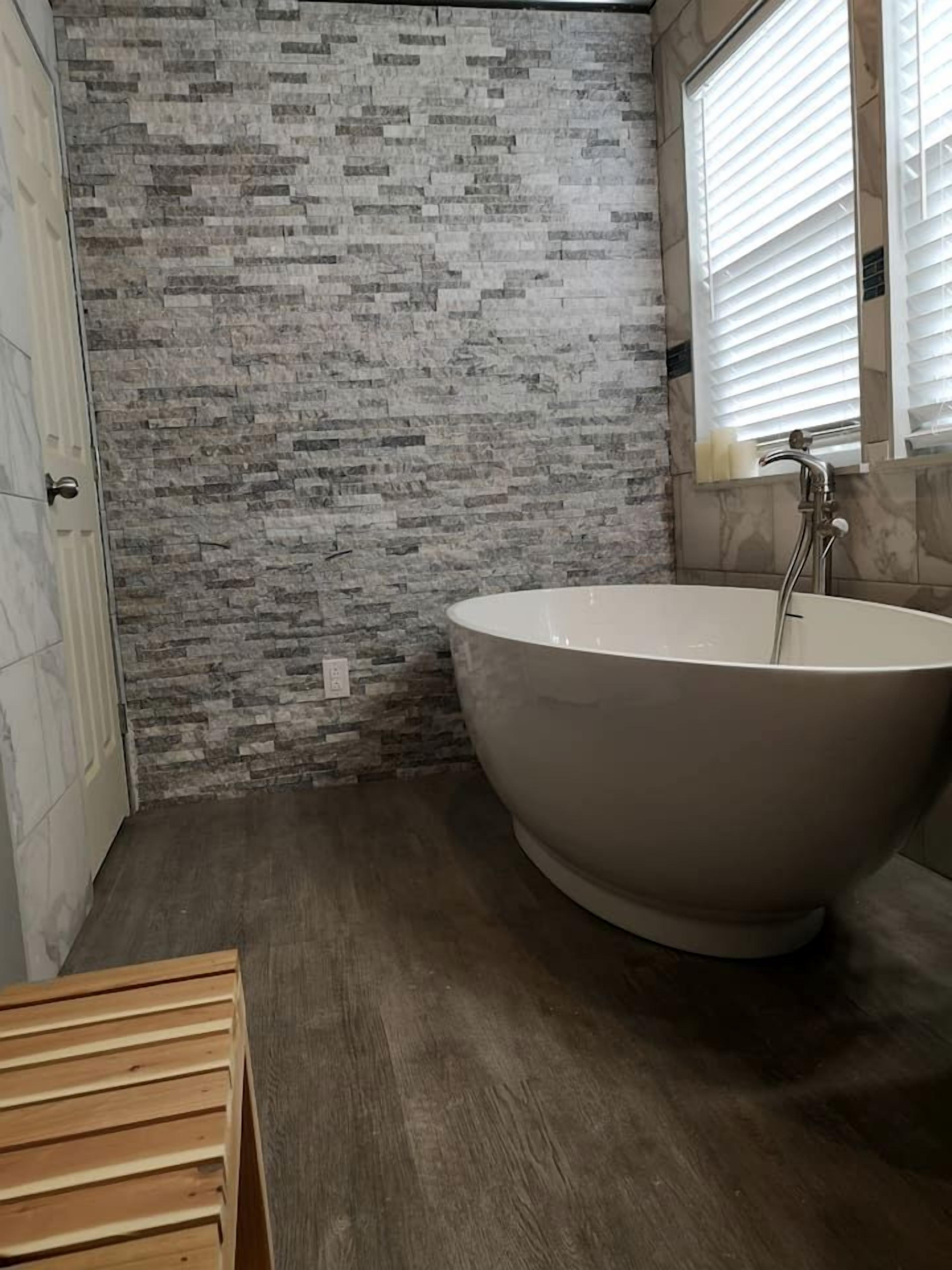 A bathroom with a freestanding bathtub, a stone wall, and a window with blinds.