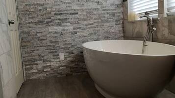 A bathroom with a freestanding bathtub, a stone wall, and a window with blinds.