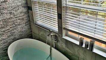 A bathroom with a freestanding bathtub, two windows with blinds, and a view of a wooden structure outside.