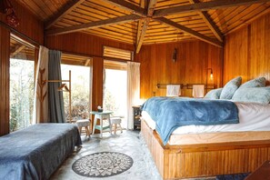 Quarto de cabana de madeira com uma cama, duas mesinhas de cabeceira, um banquinho e uma janela com cortinas.