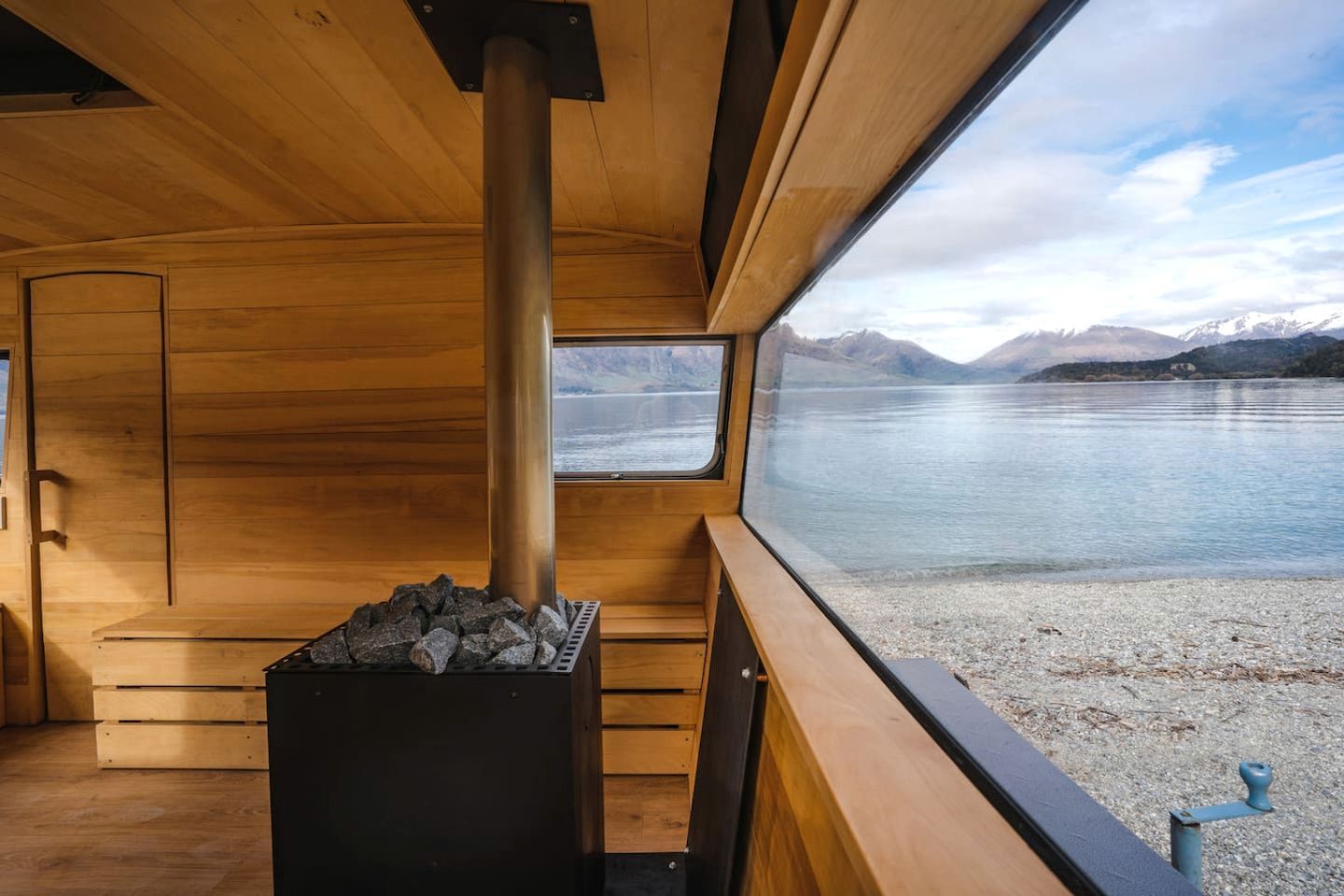 A wooden cabin interior with a fireplace, a window overlooking a lake, and a view of mountains.