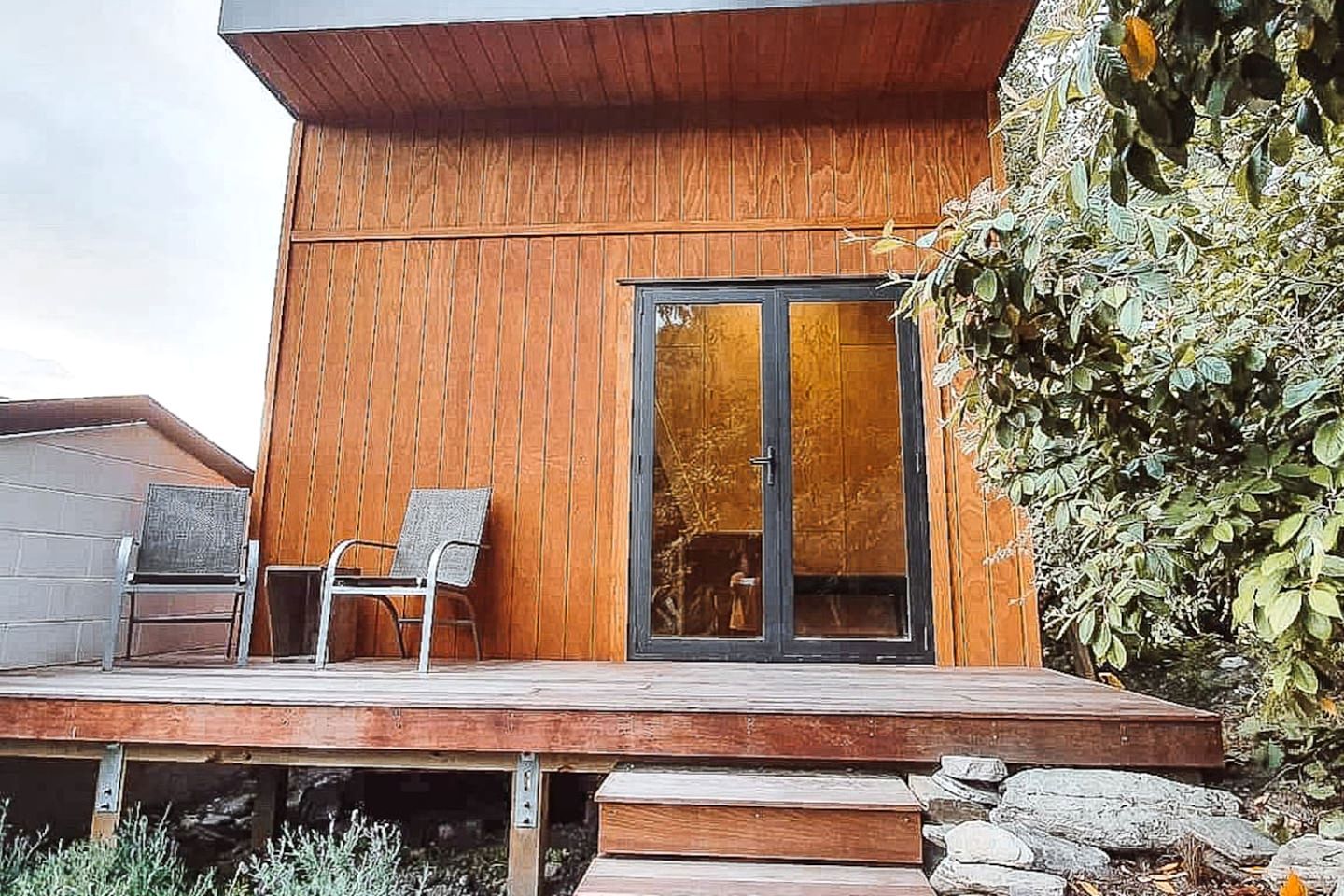 A modern building with a wooden exterior, a glass door, and a wooden deck with two chairs.
