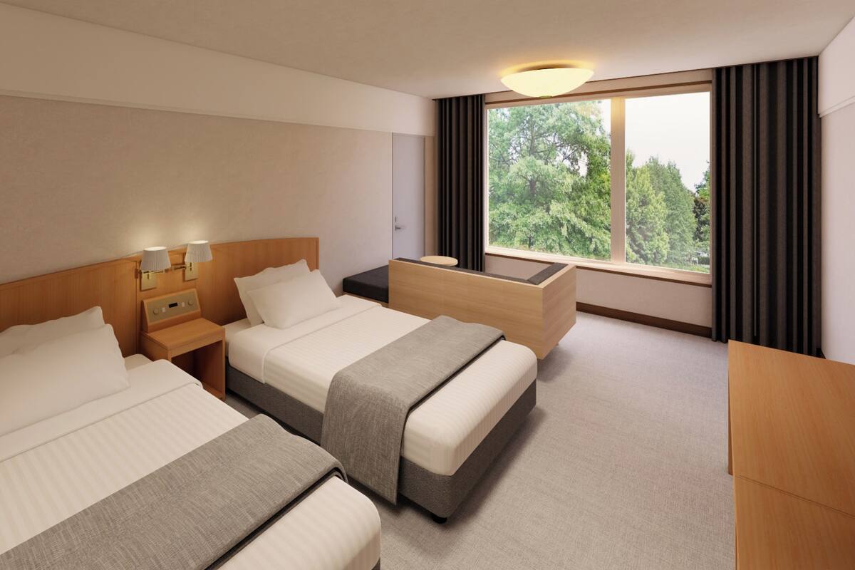 A hotel room with two beds, a bedside table, a desk, and a window with a view of trees.