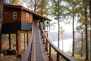 A wooden treehouse with a wooden deck leading to a balcony, surrounded by trees and a body of water.