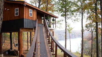 Una casa del árbol de madera con un balcón accesible mediante una terraza de madera, rodeada de árboles y una masa de agua.
