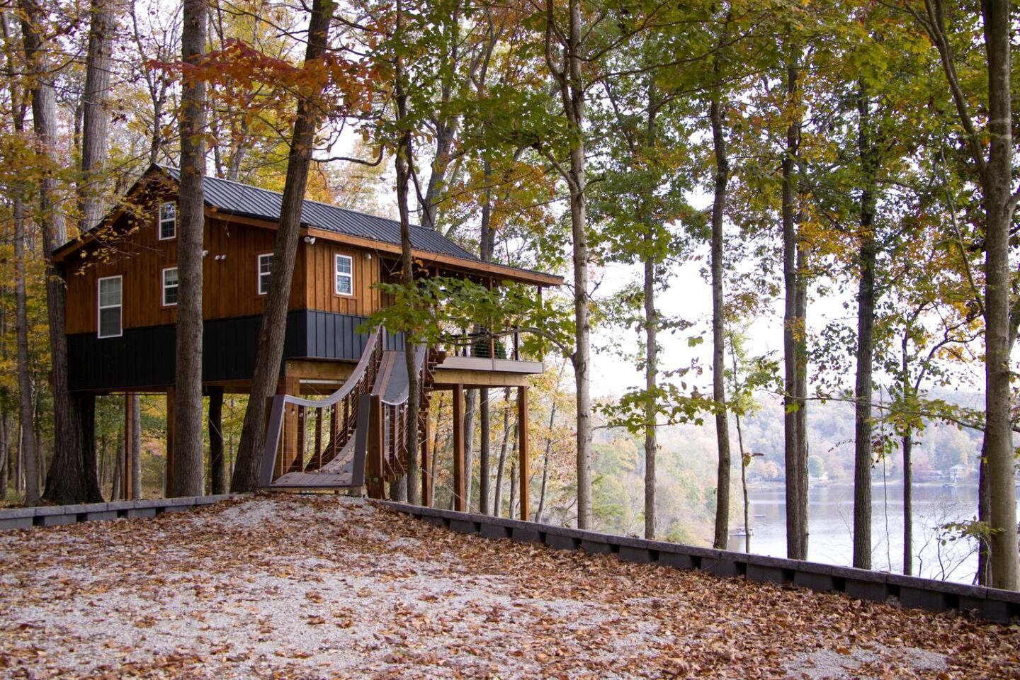 A treehouse cabin with a wooden exterior, a staircase leading up, and a view of a lake.