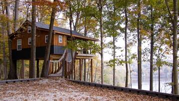 A treehouse cabin with a wooden exterior, a staircase leading up, and a view of a lake.