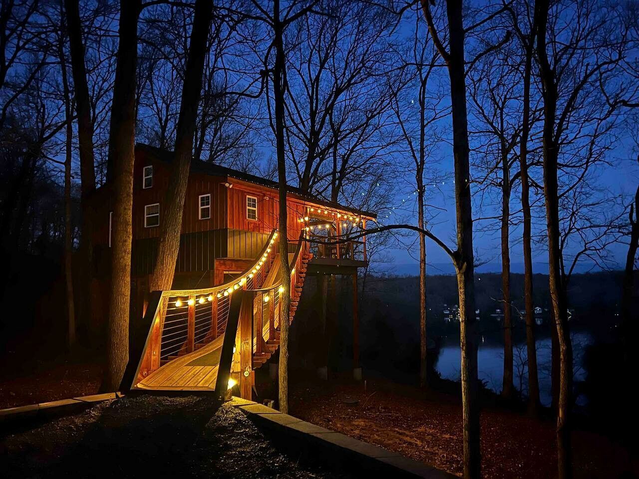 A wooden cabin with a deck and staircase, surrounded by trees and a body of water.