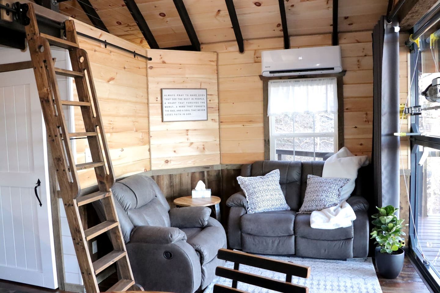 A cozy cabin interior with a wooden ceiling, a gray recliner, a small round table, and a potted plant.