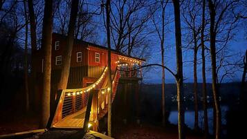 A wooden cabin with a deck and staircase, surrounded by trees and a body of water.