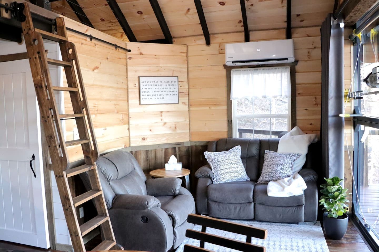 A cozy cabin interior with a wooden ceiling, a gray recliner, a small round table, and a potted plant.