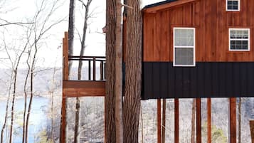Une cabane perchée dans un arbre, dotée d’une terrasse en bois et d’une partie inférieure noire, entourée d’arbres et bordée par un plan d’eau.