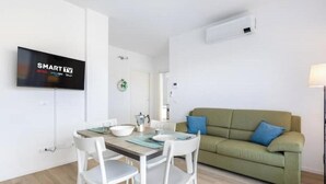 Una sala de estar moderna con una mesa de comedor, sillas blancas, un sofá verde con cojines azules y un televisor montado.