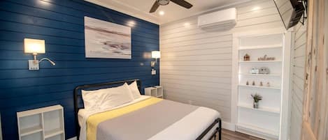 Un dormitorio con cama, mesitas de noche, ventilador de techo, equipo de aire acondicionado y estante empotrado con objetos decorativos.