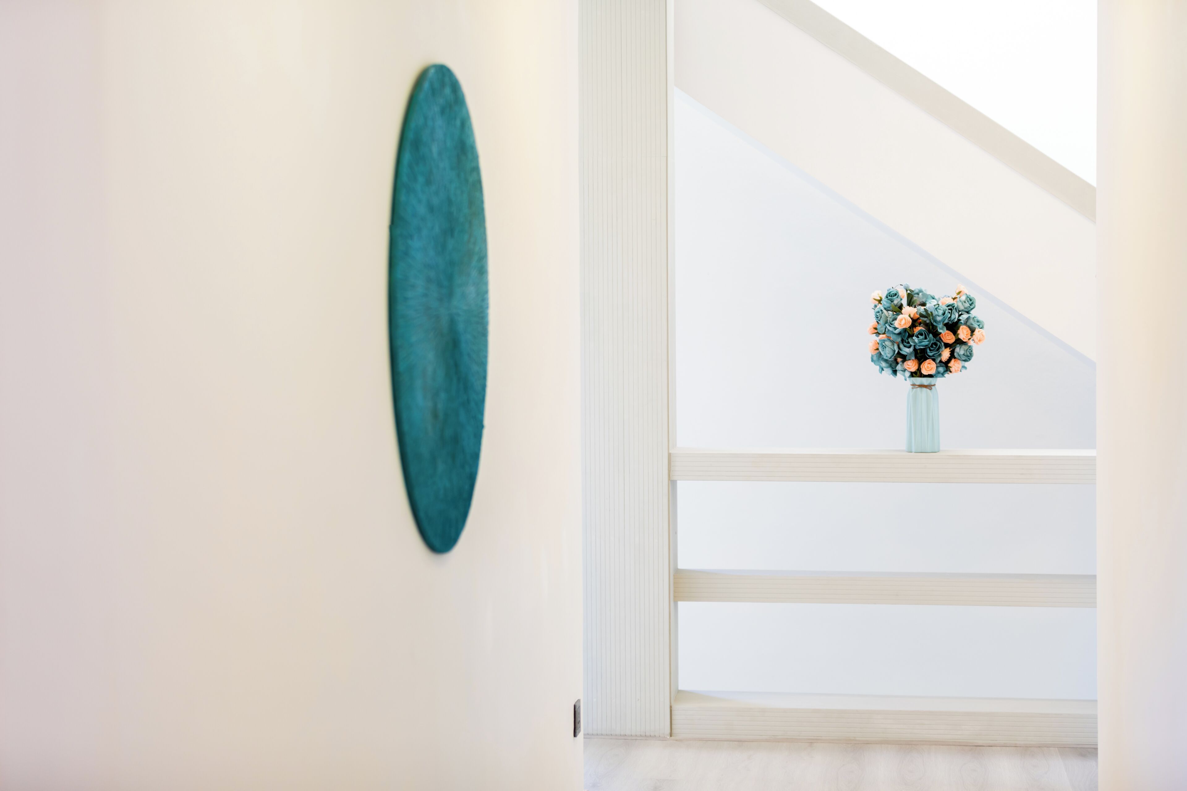 A hallway with a teal abstract painting on the wall and a vase with flowers on a window sill.