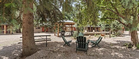 Un espacio de descanso rústico al aire libre con sillas Adirondack, una mesa de picnic y una cabaña de madera al fondo.
