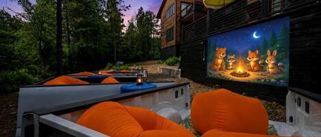 An outdoor seating area with orange cushions, a projector screen displaying animated characters, and a wooden building in the background.