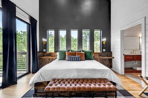 A bedroom with a large bed, a bench, and a view of trees through the windows. - Luxury Cabin with Heated Pool, Sauna & Hot Tub Retreat in Broken Bow, Oklahoma (Broken Bow)