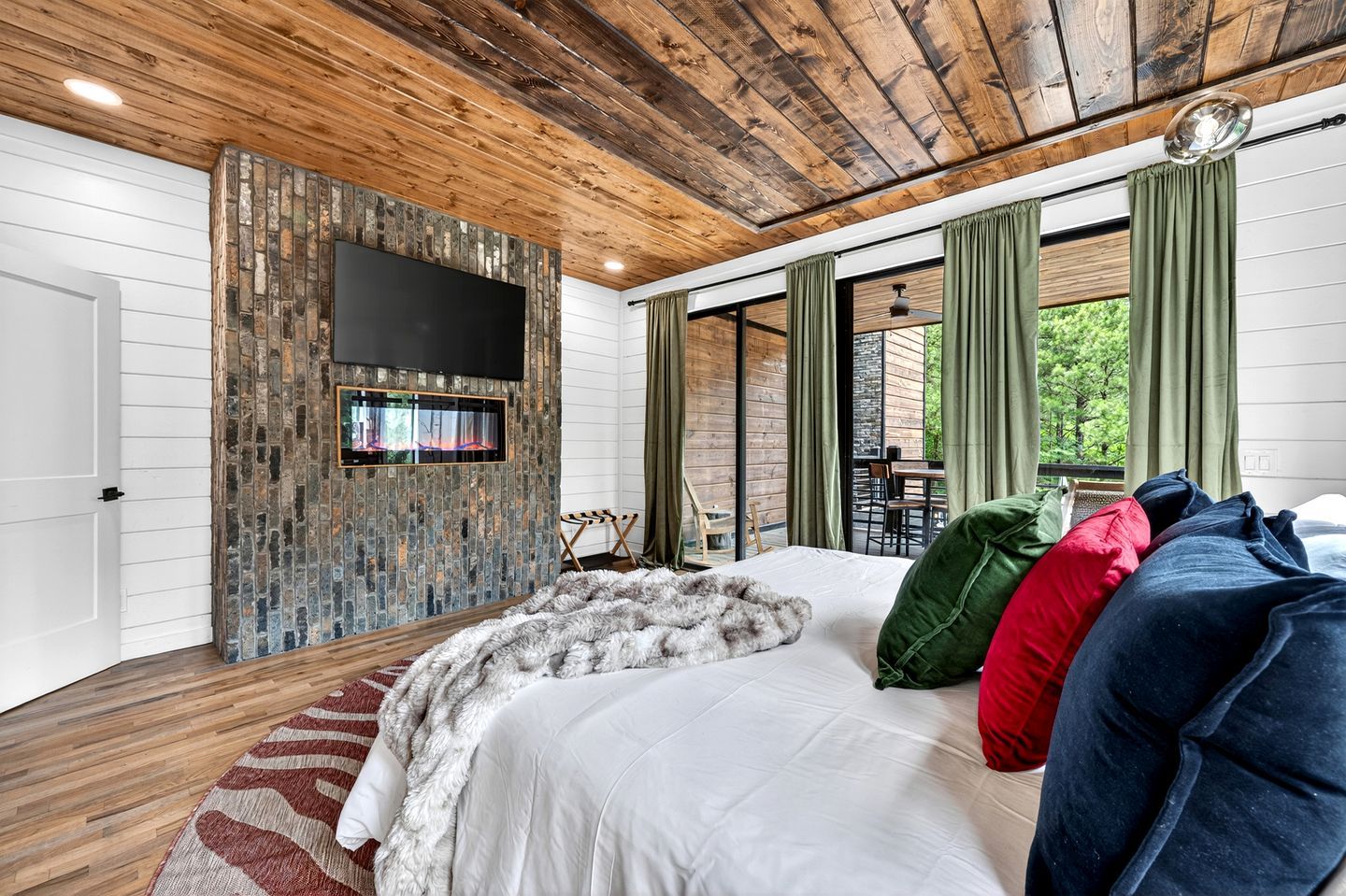 Un dormitorio con chimenea de piedra, una cama con muchos cojines, un ventanal con cortinas y vistas al exterior.