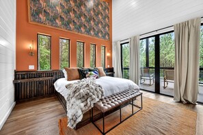A bedroom with a large bed, a bench, a view of the outdoors, and a painting on the wall. - Luxury Cabin with Heated Pool, Sauna & Hot Tub Retreat in Broken Bow, Oklahoma (Broken Bow)