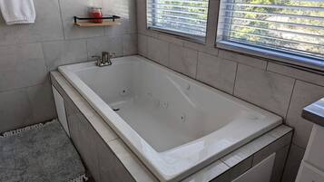 Une salle de bain moderne avec une grande baignoire, des murs carrelés, une petite étagère avec une bougie et une fenêtre avec des stores.